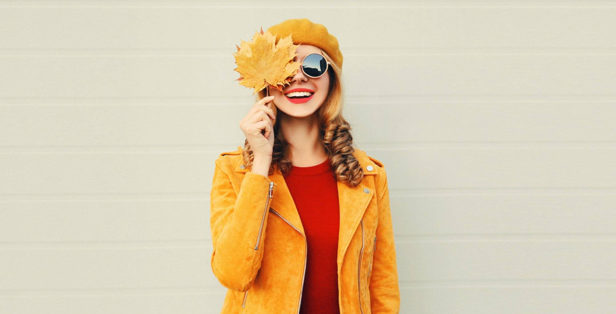 Autumn mood! happy smiling woman holding in her hands yellow maple leaves hiding her eye over gray wall background Autumn mood! happy smiling woman holding in her hands yellow maple leaves hiding her eye over gray wall background
