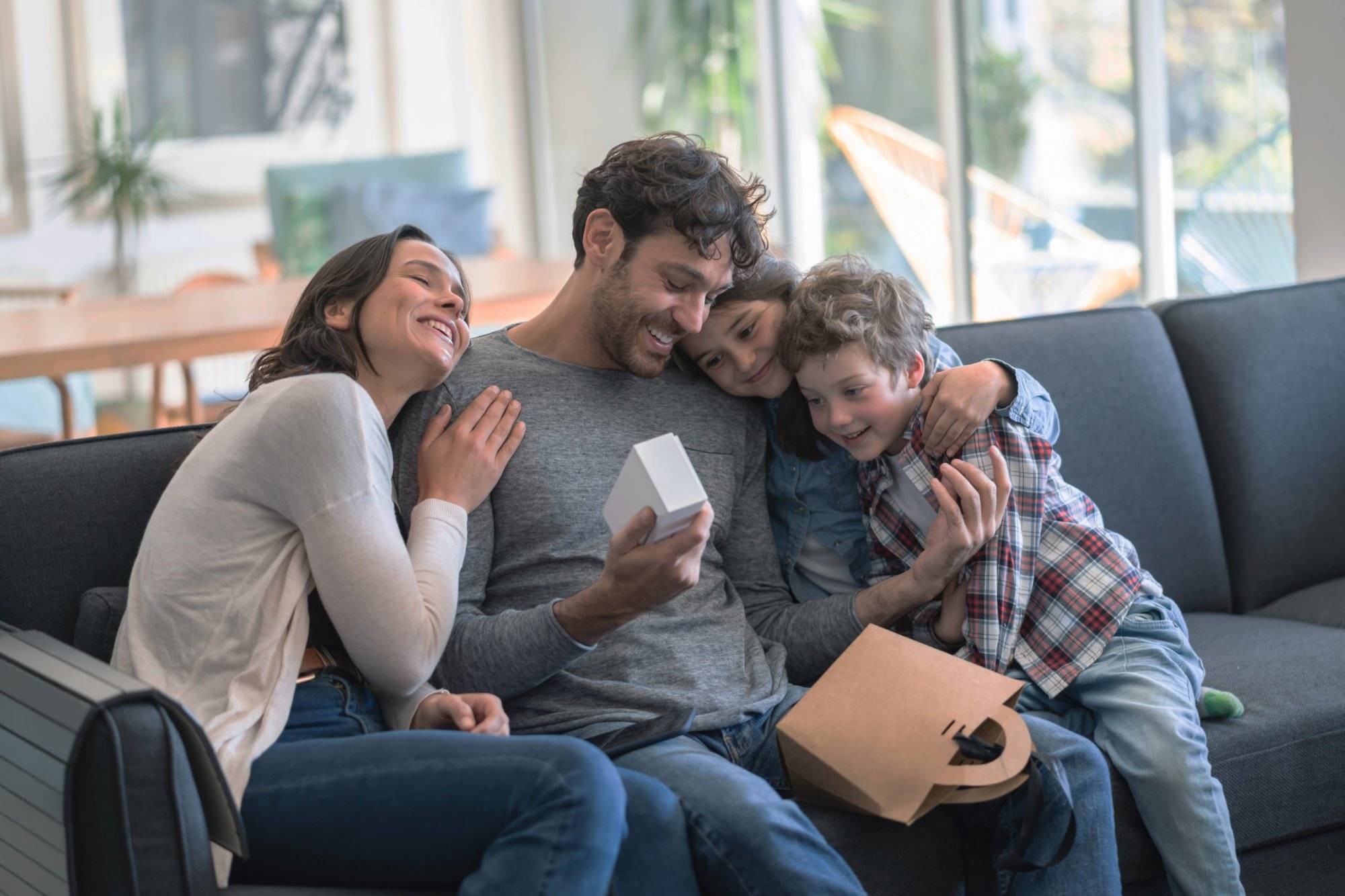 Loving mother and kids celebrating father;s day hugging daddy while he opnes a present all smiling Loving mother and kids celebrating father;s day hugging daddy while he opnes a present all smiling