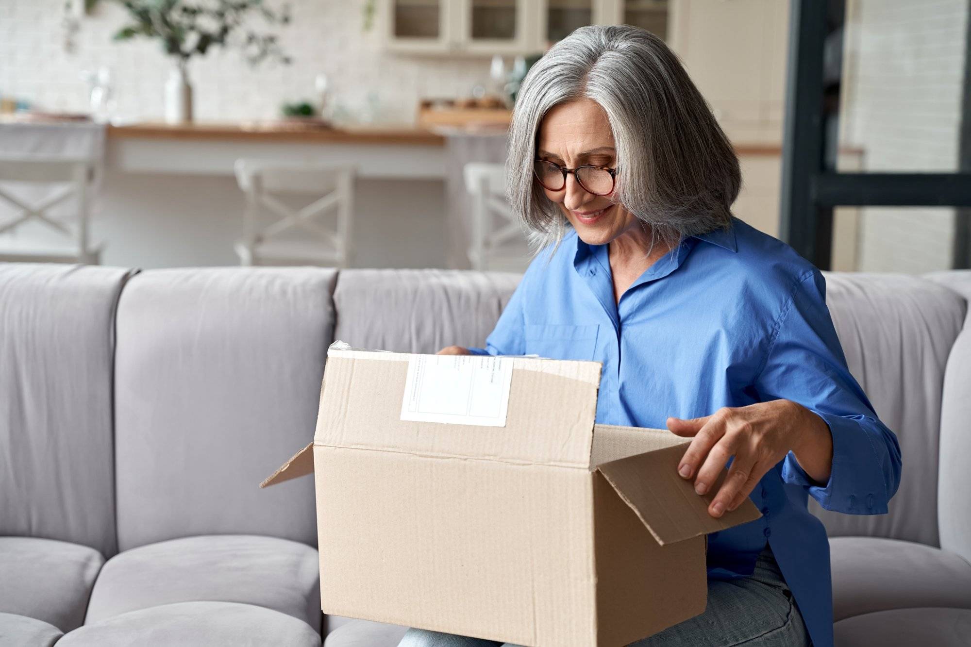 Smiling older adult mature woman customer unpacking parcel concept sitting at home on couch. Happy senior middle aged lady opening online store order receiving gift in postal delivery shipping box.